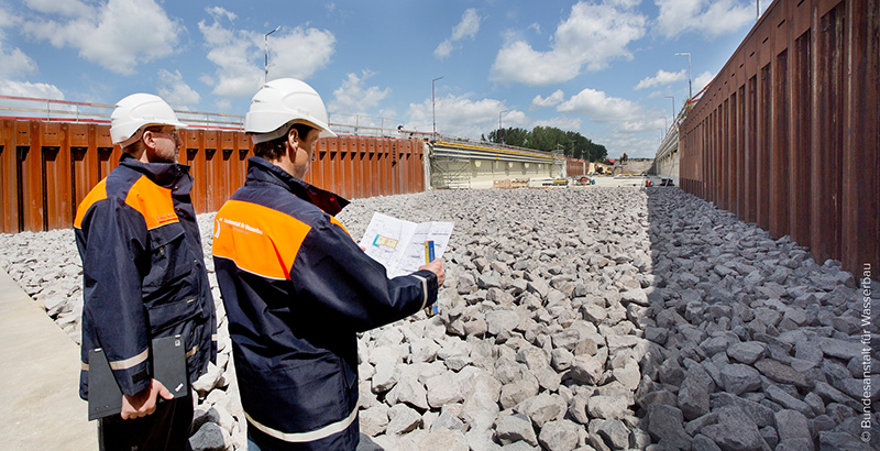 Two engineers with white safety helmets and blue-orange warning jackets, one holding a construction plan in hand, are inspecting a drained lock.
