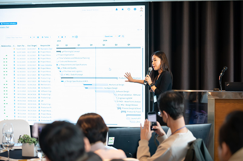 A young woman presents a Gantt chart in CONTACT Elements on a large screen in front of an audience.