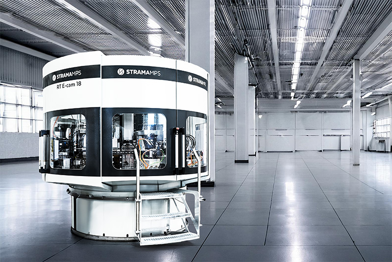 A black and white rotary transfer machine with the inscription "STRAMA MPS" in a gray factory hall.